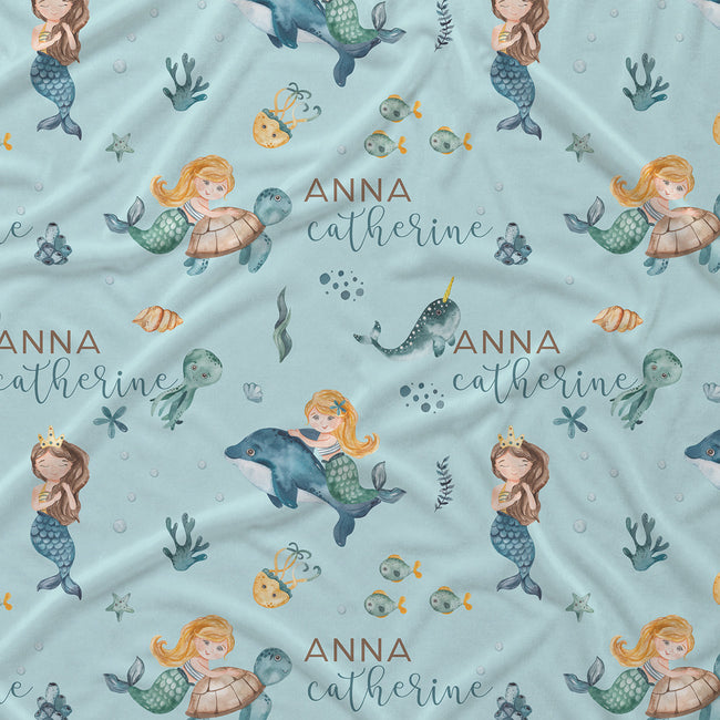 Personalized baby bedding or fabric featuring a light blue background with a whimsical mermaid and sea creature print, including the custom name 'ANNA catherine' in brown and gray script.