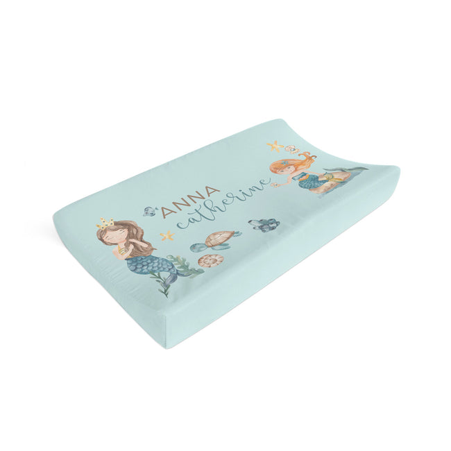 A contoured baby changing pad cover in light blue, personalized with the name 'ANNA catherine' and decorated with cute mermaid and sea turtle illustrations, shown on a plain white background.
