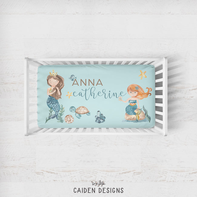 A personalized baby bedding set displayed on a white wooden crib, featuring a light blue muslin blanket and matching pillowcase with watercolor mermaid illustrations and the name 'Anna Catherine'.