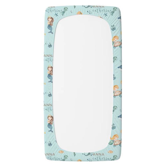 A personalized twin fitted sheet with a light blue mermaid-themed border. The border features cartoon illustrations of mermaids, sea creatures, and the names 'Anna' and 'Catherine' printed in a gold script font.