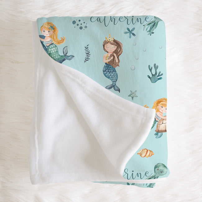 A personalized baby blanket with a white plush reverse side and a light blue printed front featuring mermaids, sea creatures, and the name 'Catherine'.