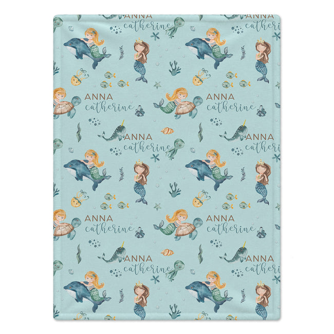 A personalized baby blanket featuring a light blue mermaid and dolphin pattern with the name 'Anna Catherine' printed in gold script.