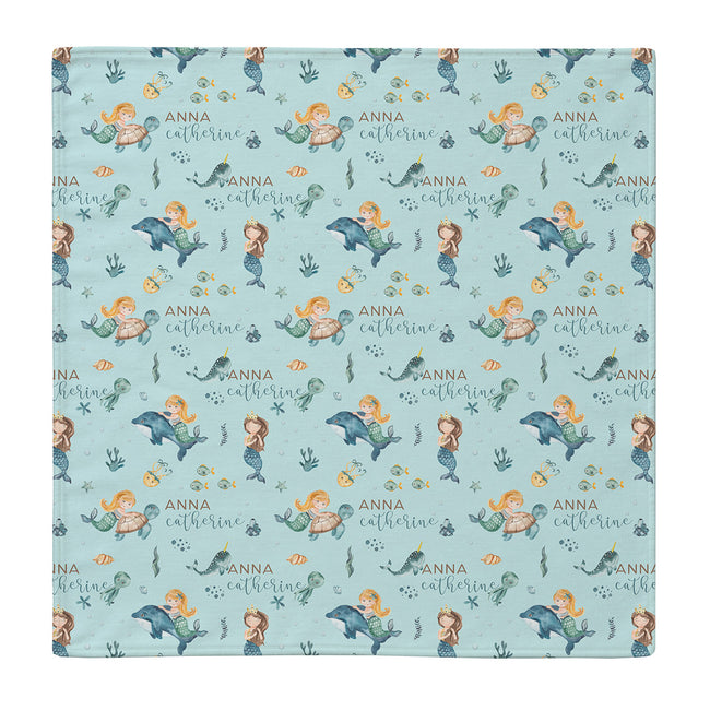 A square fabric item, likely a baby blanket or cloth, featuring a repeating pattern of mermaids, dolphins, and the personalized name 'anna catherine' on a light blue background.