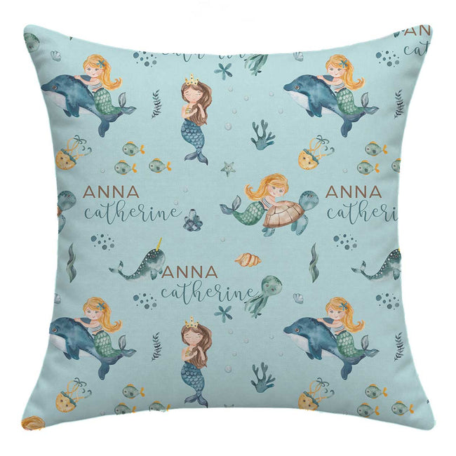 A square throw pillow with a light blue mermaid and underwater theme print, featuring the personalized name 'ANNA catherine' in brown and teal script. The pattern includes mermaids, dolphins, fish, and sea plants on a solid light blue background.