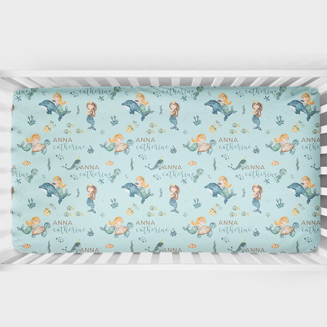 A crib mattress fitted sheet with a whimsical mermaid and dolphin pattern on a light blue background, personalized with the names 'ANNA' and 'catherine' in script.