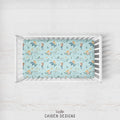 A crib mattress fitted sheet with a mermaid and sea creature pattern in light blue, displayed inside a white wooden crib on a white wood floor. The brand name 'CAIDEN DESIGNS' is printed at the bottom.