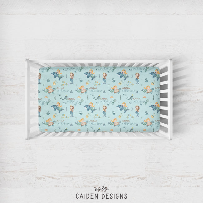 A crib mattress fitted sheet with a mermaid and sea creature pattern in light blue, displayed inside a white wooden crib on a white wood floor. The brand name 'CAIDEN DESIGNS' is printed at the bottom.