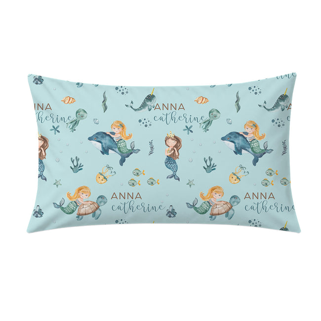A rectangular pillow with a light blue mermaid-themed pattern featuring cartoon mermaids, dolphins, fish, and the name 'Anna Catherine' printed repeatedly, shown on a plain white background.