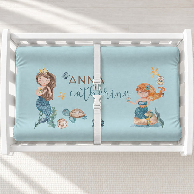 A white changing table with a personalized changing pad cover featuring watercolor mermaid illustrations.