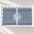 A changing pad cover with a monogrammed design, displayed inside a white wooden baby crib. The blanket features a repeating 'BECKETT' brand pattern in light blue-gray on a medium blue-gray background, with a large circular monogram 'BKH' in the center.
