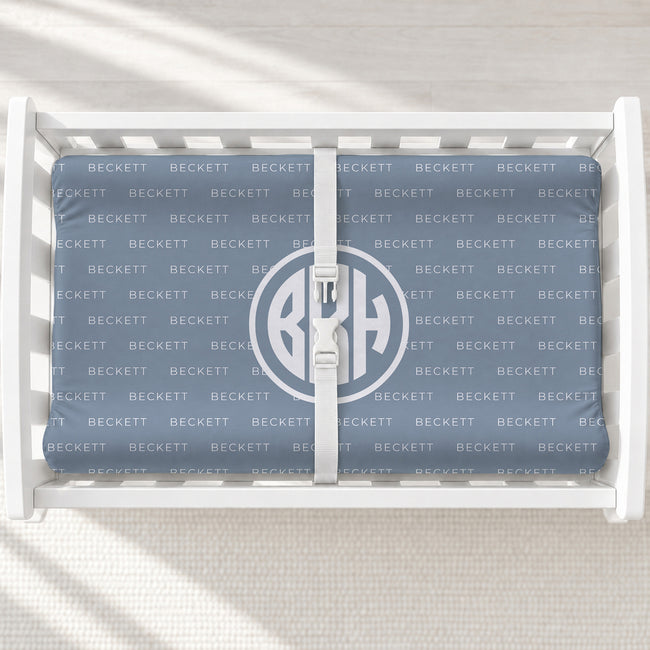 A changing pad cover with a monogrammed design, displayed inside a white wooden baby crib. The blanket features a repeating 'BECKETT' brand pattern in light blue-gray on a medium blue-gray background, with a large circular monogram 'BKH' in the center.