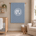 A personalized baby blanket featuring a monogrammed 'BMJ' in a white circle over a repeating light blue and white 'Brooks' pattern, displayed in a styled nursery room next to a wooden crib and a beige armchair.