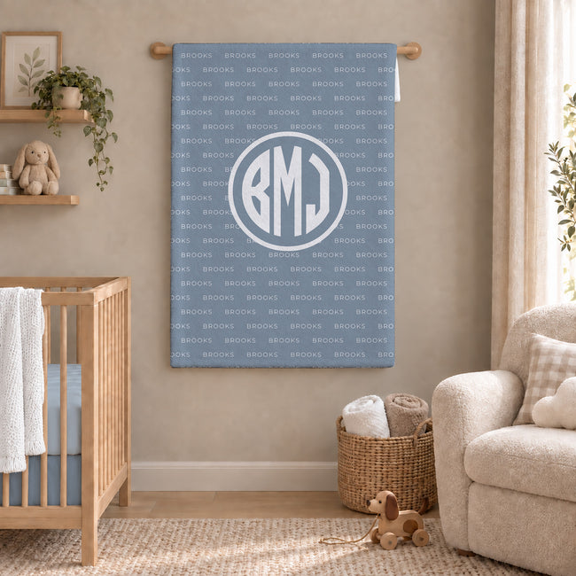A personalized baby blanket featuring a monogrammed 'BMJ' in a white circle over a repeating light blue and white 'Brooks' pattern, displayed in a styled nursery room next to a wooden crib and a beige armchair.