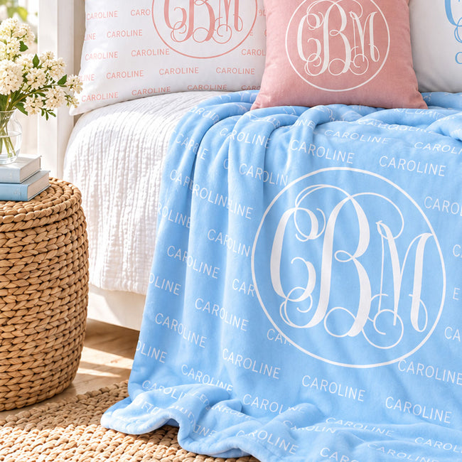 Light blue personalized blanket with the repeated name Caroline and a large white circular monogram, draped over a white bed with coordinating pink and white pillows.