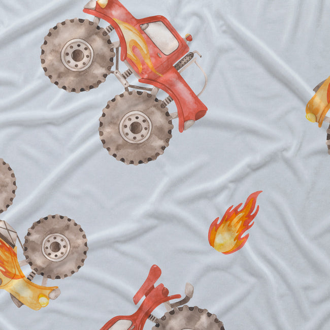 Close-up of watercolor monster trucks with oversized taupe tires and orange flame accents on pale blue fabric.