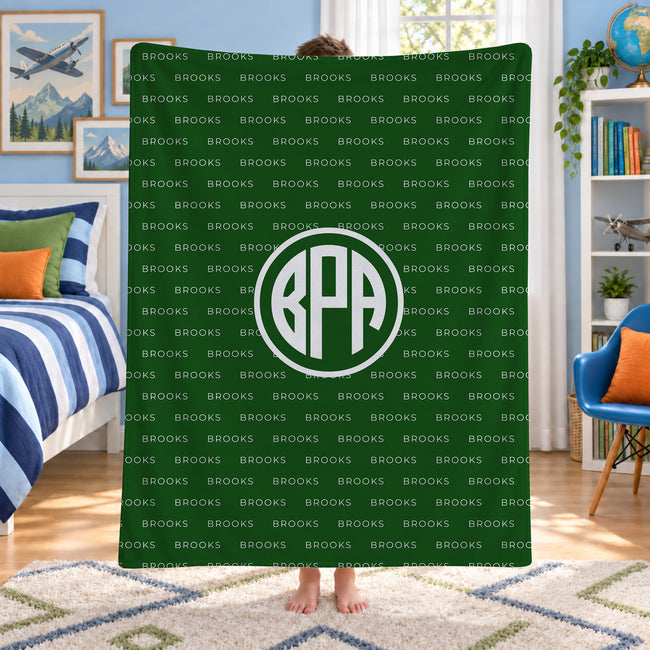 A person standing in a bedroom holding up a dark green blanket or throw featuring a repeating 'BROOKS' wordmark pattern and a large circular 'BPA' monogram in the center.