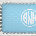 A light blue baby crib bedding set featuring a repeating 'BECKETT' monogram pattern and a large custom white circular monogram with the initials 'BWH' on the side.