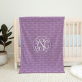 A personalized purple baby blanket with a repeating 'PALMER' monogram pattern and a large central 'PSC' monogram, draped over a white wooden crib in a nursery setting.