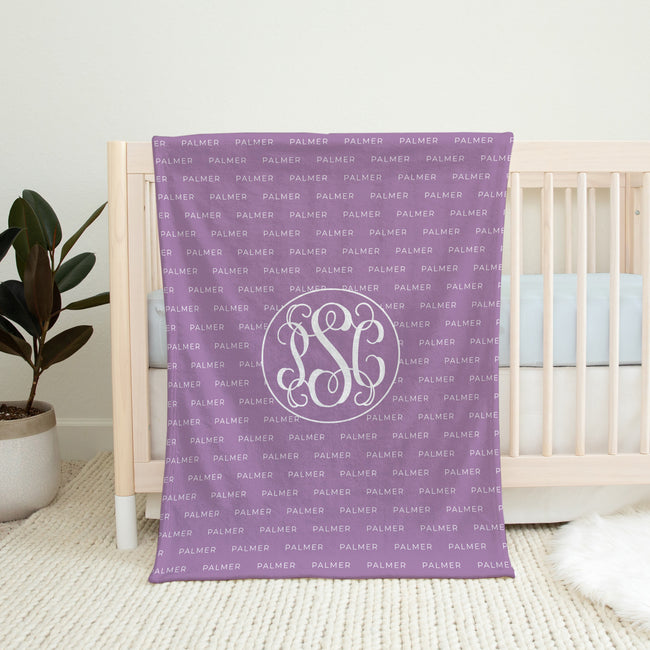 A personalized purple baby blanket with a repeating 'PALMER' monogram pattern and a large central 'PSC' monogram, draped over a white wooden crib in a nursery setting.