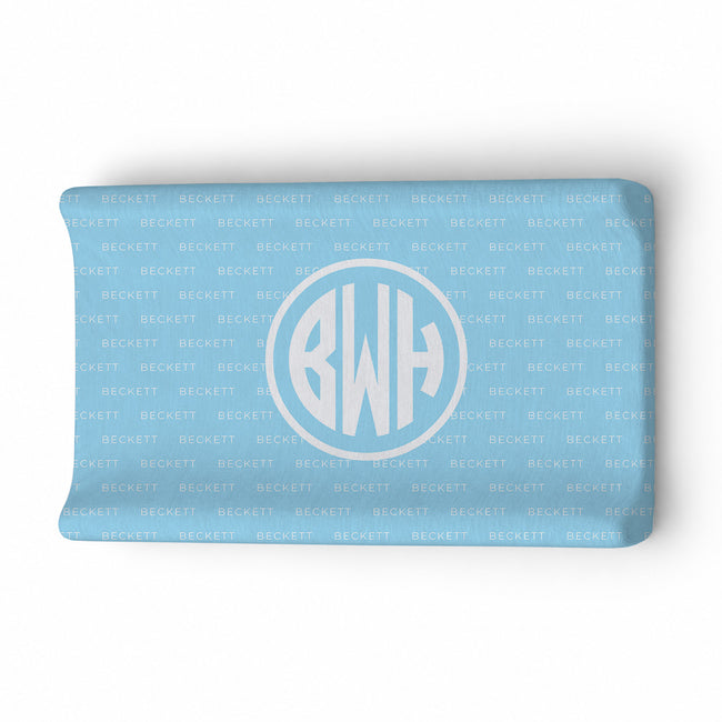A light blue changing pad cover featuring a repeating 'BECKETT' pattern and a large central circular monogram with the initials 'BWH' in white.