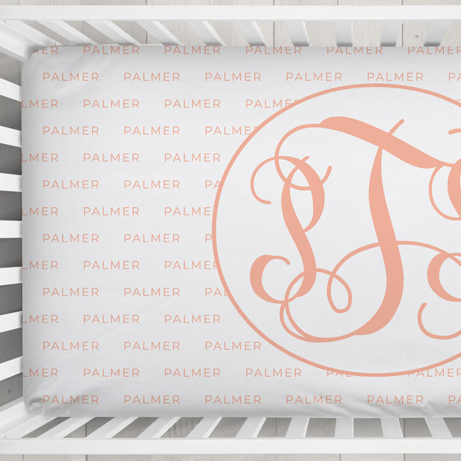 A close-up of a white crib sheet featuring a large peach-pink monogram letter 'S' inside a circle, with the brand name 'PALMER' repeated faintly in peach-pink text across the background.