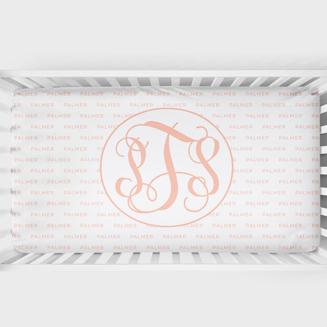 A crib sheet featuring a large peach monogram design in the center with the initials 'PS' in an elegant script. The background of the crib sheet is covered in a repeating light pink 'PALMER' brand watermark pattern.