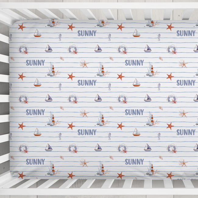 A crib mattress fitted sheet with a nautical theme, featuring the word 'SUNNY' in blue, sailboats, life preservers, seahorses, and orange starfish on a white background with light blue horizontal stripes.