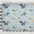 A crib mattress fitted sheet with an underwater-themed pattern featuring whales, dolphins, sea turtles, jellyfish, and the name 'grace' in script, shown inside a white wooden crib.