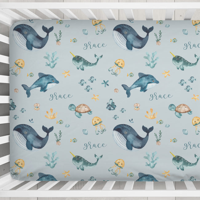 A crib mattress fitted sheet with an underwater-themed pattern featuring whales, dolphins, sea turtles, jellyfish, and the name 'grace' in script, shown inside a white wooden crib.