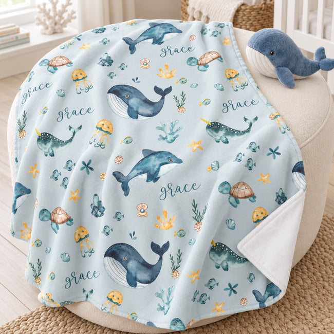 Personalized Sea Animal Baby Blanket. Whales, dolphins, turtles, narwhals, and other ocean friends surround a custom name on a soft blue-grey background.