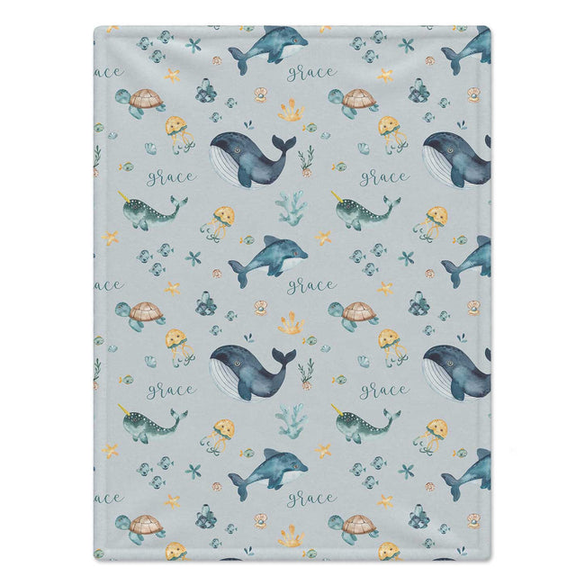 Personalized Sea Animal Baby Blanket. Whales, dolphins, turtles, narwhals, and other ocean friends surround a custom name on a soft blue-grey background.