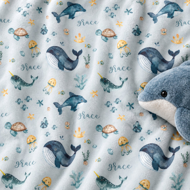 A close-up of a plush blanket or fabric featuring an underwater print with whales, jellyfish, turtles, narwhals, and the word Grace repeated in script. A blue-and-white plush whale toy is partially visible on the right side.