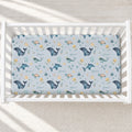 A fitted crib mattress with an ocean-themed print featuring whales, sea turtles, dolphins, and the word 'Grace' in blue, teal, yellow, and brown on a light blue background, shown inside a white wooden crib.