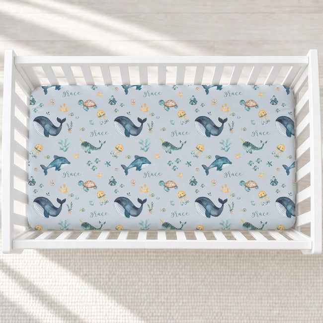 A fitted crib mattress with an ocean-themed print featuring whales, sea turtles, dolphins, and the word 'Grace' in blue, teal, yellow, and brown on a light blue background, shown inside a white wooden crib.