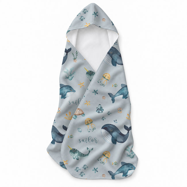 Light blue hooded towel with whales, dolphins, narwhals, turtles, jellyfish, shells, starfish, and the name Sailor in script.