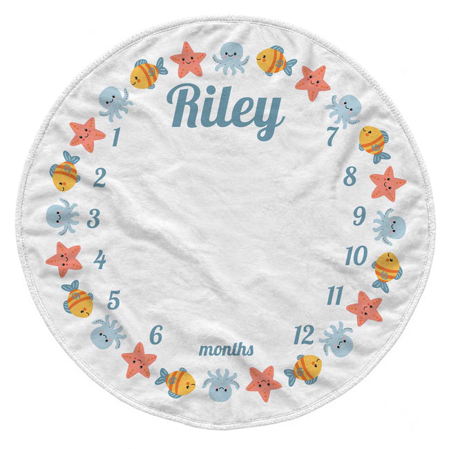 A circular white baby milestone chart blanket featuring colorful cartoon sea creatures and the name 'Riley' in blue script, with numbered months from 1 to 12 around the border.