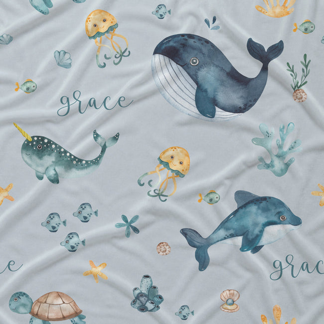 Light gray fabric with a watercolor-style ocean print featuring a blue whale, blue dolphin, teal narwhal, yellow jellyfish, seashells, coral, and the word Grace repeated in script.