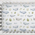 A crib mattress fitted sheet with a repeating pattern of blue-gray whales, narwhals, green leaves, and the name 'RILEY' printed in blue, shown inside a white wooden crib.