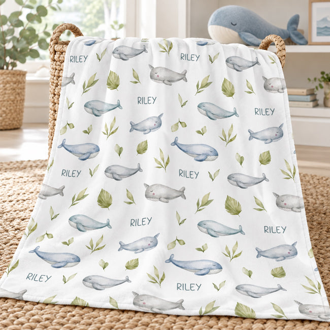 A white baby blanket draped over a woven basket, featuring a repeating pattern of light blue whales, green leaves, and the names 'Riley' printed in blue. The blanket is styled in a bright, modern nursery setting.