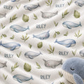 A close-up of a soft blanket or fabric print featuring watercolor-style blue and gray whales, green leaves, and the name 'RILEY' repeated on an off-white background, with a plush whale toy partially visible in the lower right corner.