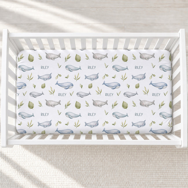 A white crib mattress fitted sheet with a repeating pattern of blue-gray whales, green leaves, and the name 'RILEY' printed in dark lettering, shown inside a white crib on a light textured floor.