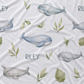 White fabric with a watercolor-style print of gray whales, narwhals, green leaves, and the name 'RILEY' repeated across the surface.