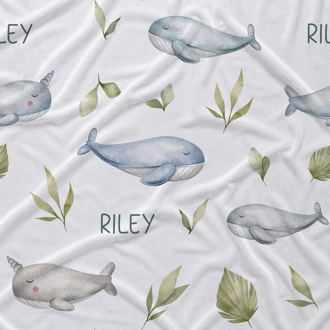 White fabric with a watercolor-style print of gray whales, narwhals, green leaves, and the name 'RILEY' repeated across the surface.