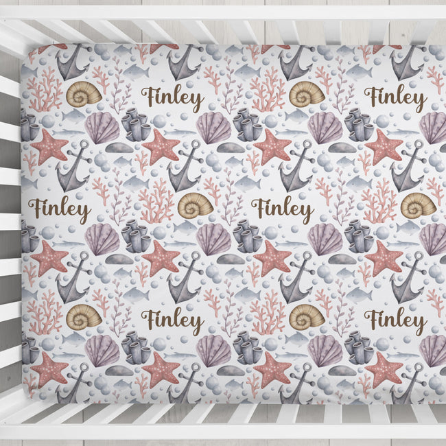 A crib mattress cover with a personalized nautical pattern featuring the name 'Finley' in brown script, decorated with seashells, starfish, anchors, and coral on a white background.