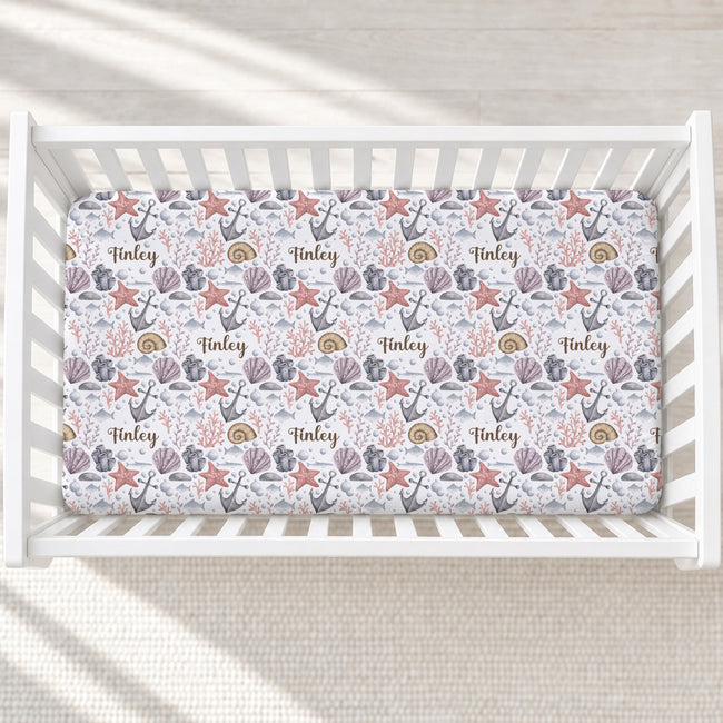 A baby crib mattress fitted sheet with a nautical pattern of starfish, anchors, shells, and the name 'Finley' repeated, shown inside a white wooden crib.
