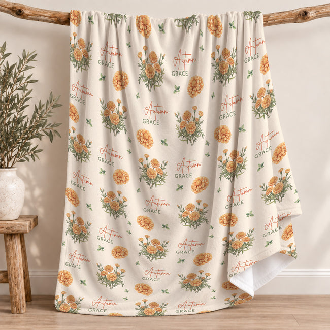 Cream blanket with golden-orange marigold clusters, scattered green leaves, and the name Autumn Grace in orange and green.