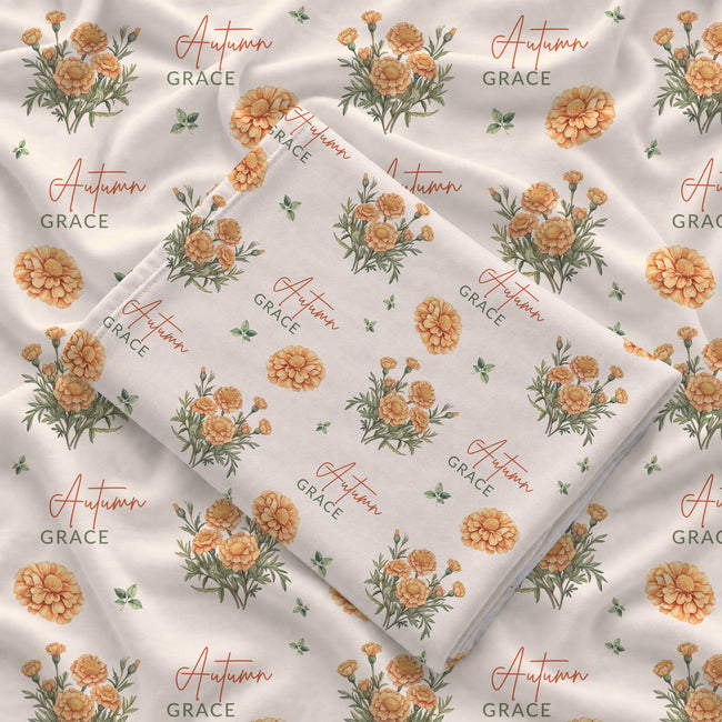 Cream swaddle blanket with golden-orange marigold clusters, green leaves, and the name Autumn Grace in orange and green.