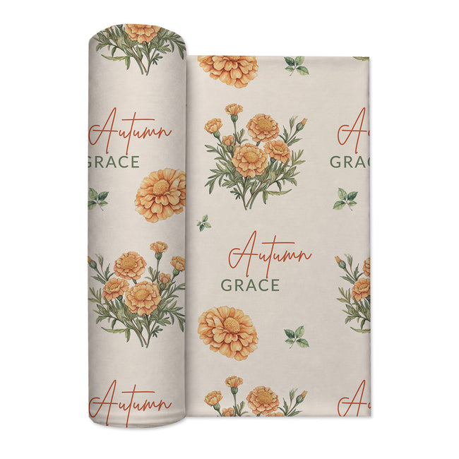 Cream swaddle blanket with golden-orange marigold clusters, green leaves, and the name Autumn Grace in orange and green.