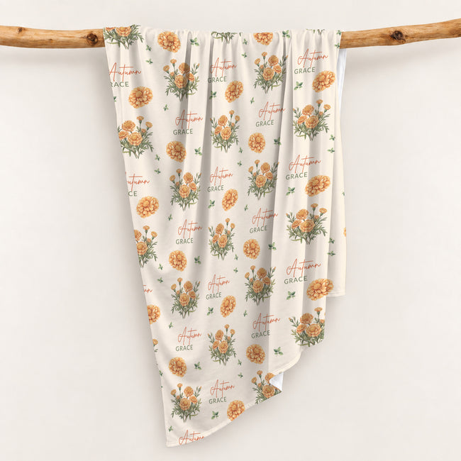 Cream swaddle blanket with golden-orange marigold clusters, green leaves, and the name Autumn Grace in orange and green.
