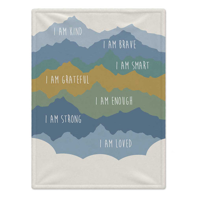 A personalized blanket featuring a layered mountain design in shades of blue, green, and mustard yellow, with white hand-lettered text reading positive affirmations like 'I AM KIND', 'I AM BRAVE', and 'I AM LOVED'.